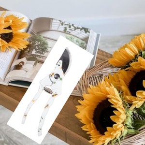 May include: White gardening shears with a bee and leaf design. The shears are on a wooden table with sunflowers in the background.