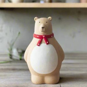 May include: A ceramic bear ornament with a beige body, a white belly, and a red scarf with white spots. The bear has a friendly expression and is standing upright. The ornament is a decorative item.