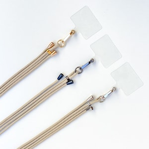 K&ouml;nnte beinhalten: Drei beigefarbene Lanyards mit verschiedenen farbigen Metallverschl&uuml;ssen und -spitzen. Die Lanyards h&auml;ngen an durchsichtigen Plastik-Etiketten.