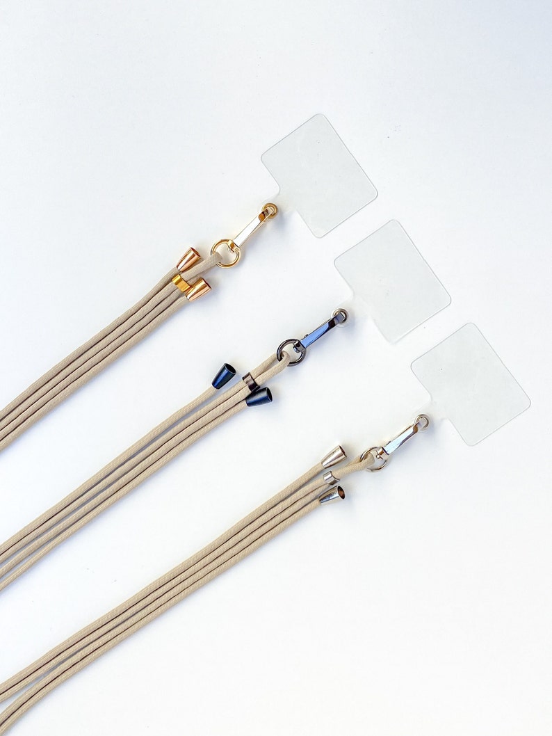 K&ouml;nnte beinhalten: Drei beigefarbene Lanyards mit verschiedenen farbigen Metallverschl&uuml;ssen und -spitzen. Die Lanyards h&auml;ngen an durchsichtigen Plastik-Etiketten.