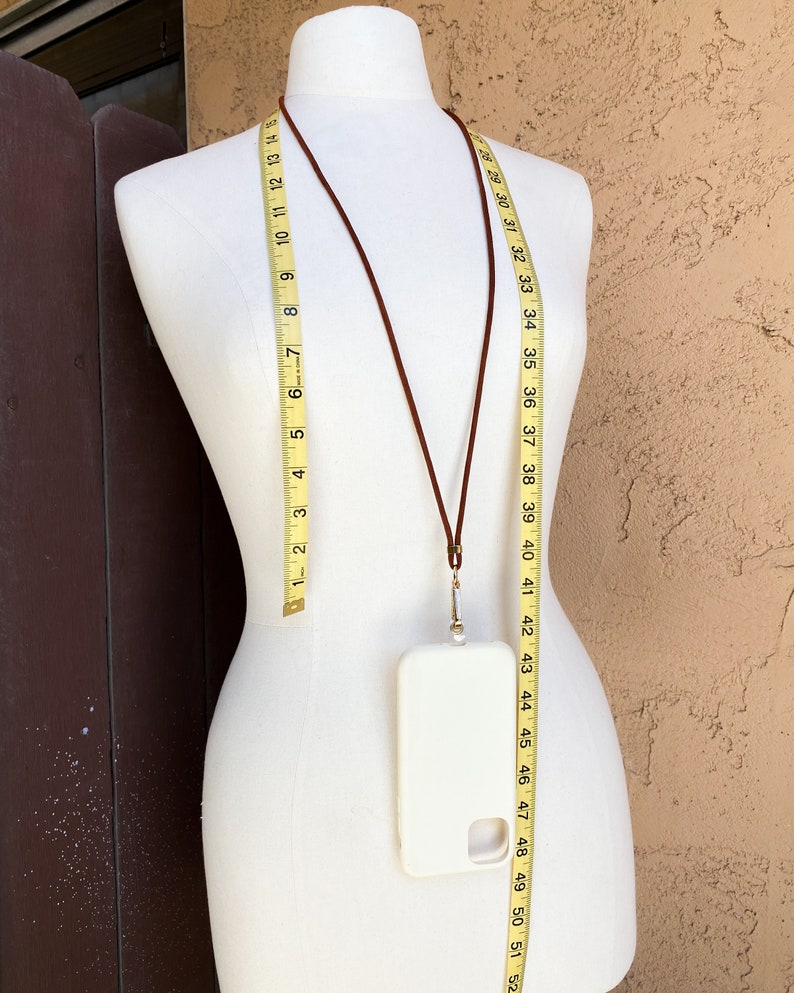 K&ouml;nnte beinhalten: Ein braunes Leder-Handyband mit einem goldenen Verschluss und einer wei&szlig;en Handyh&uuml;lle. Das Band h&auml;ngt an einer wei&szlig;en Puppe.