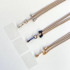 K&ouml;nnte beinhalten: Drei beigefarbene Lanyards mit verschiedenen Metallverschl&uuml;ssen und dekorativen Enden. Die Lanyards liegen auf einer wei&szlig;en Oberfl&auml;che.