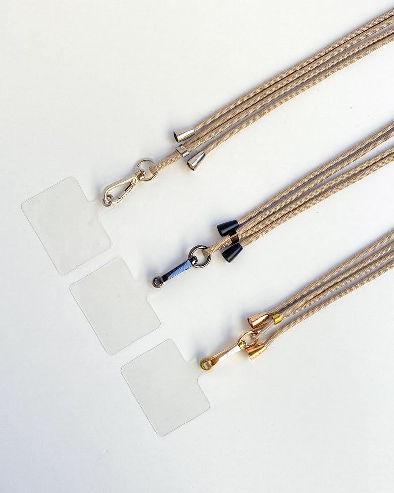 K&ouml;nnte beinhalten: Drei beigefarbene Lanyards mit verschiedenen Metallverschl&uuml;ssen und dekorativen Enden. Die Lanyards liegen auf einer wei&szlig;en Oberfl&auml;che.