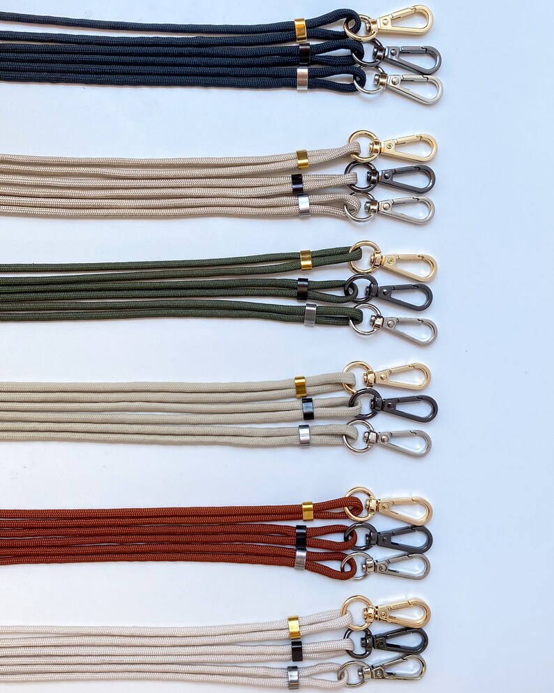 K&ouml;nnte beinhalten: F&uuml;nf S&auml;tze von Lanyards mit goldenen und schwarzen Akzenten. Jedes Lanyard hat zwei schwarze Metallverschl&uuml;sse und einen goldenen Ring. Die Lanyards sind in den Farben Schwarz, Beige, Gr&uuml;n, Braun und Wei&szlig;.