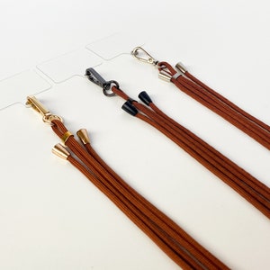 K&ouml;nnte beinhalten: Drei braune Kordel-Lanyards mit goldenen und silbernen Verschl&uuml;ssen und schwarzen und goldenen Endkappen.