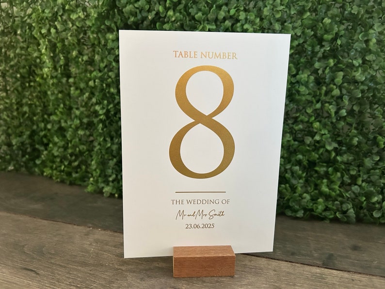 May include: White table number card with gold foil lettering. The card reads "Table Number 8" and "The Wedding of Mr. and Mrs. Smith 23.06.2025".