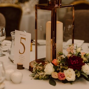 May include: A white card with gold lettering reading "Table Number 5" sits on a wooden base next to a clear glass pitcher. A copper lantern with a white candle inside sits on a wooden slice next to a bouquet of white, pink, and burgundy flowers.