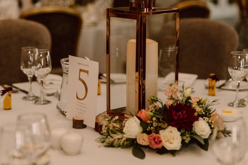 May include: A white card with gold lettering reading "Table Number 5" sits on a wooden base next to a clear glass pitcher. A copper lantern with a white candle inside sits on a wooden slice next to a bouquet of white, pink, and burgundy flowers.