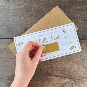 May include: A white scratch-off card with gold text that reads "We have booked for all of us, a little Treat." The card is held over a brown envelope. A coin is scratching the gold-colored area to reveal the hidden message.