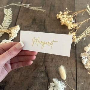 May include: White place card with the name "Margaret" written in gold script.