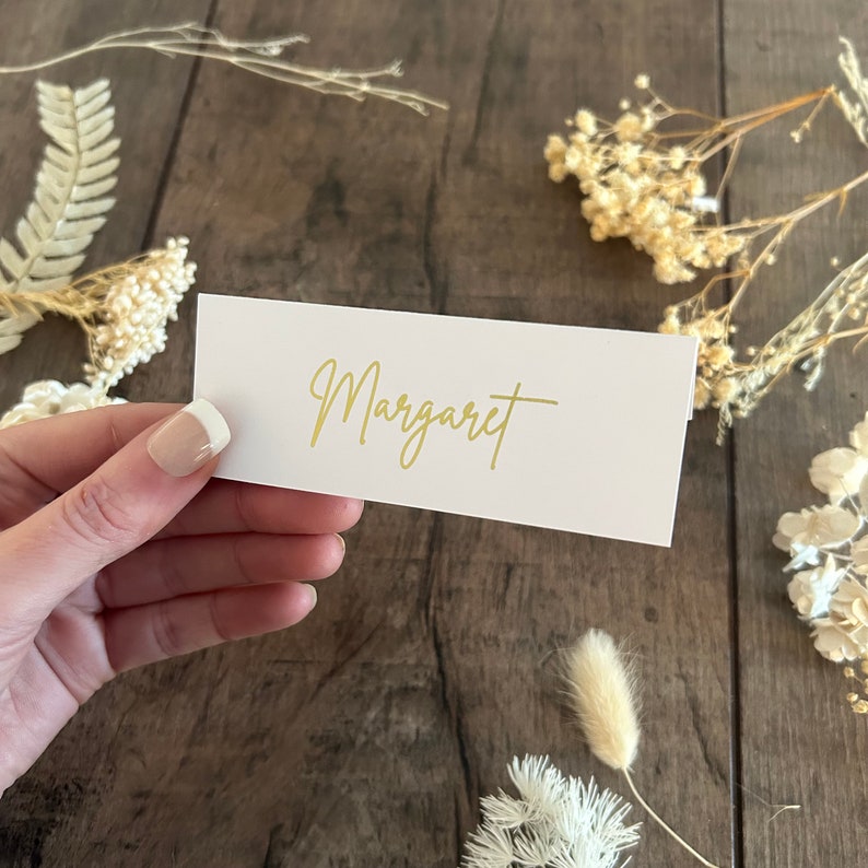 May include: White place card with the name "Margaret" written in gold script.