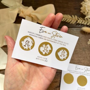 May include: A white card with gold detailing, featuring the names "Eva and Steven." The card displays three gold circles with a wine glass and heart design. Text reads "Match 3 symbols and enjoy a free drink on us!" and "Good luck! 26th June 2027."