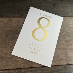 May include: White card with gold foil lettering. The card says "Table Number 8" and "The Wedding of Mr. and Mrs. Smith 23.06.2025".