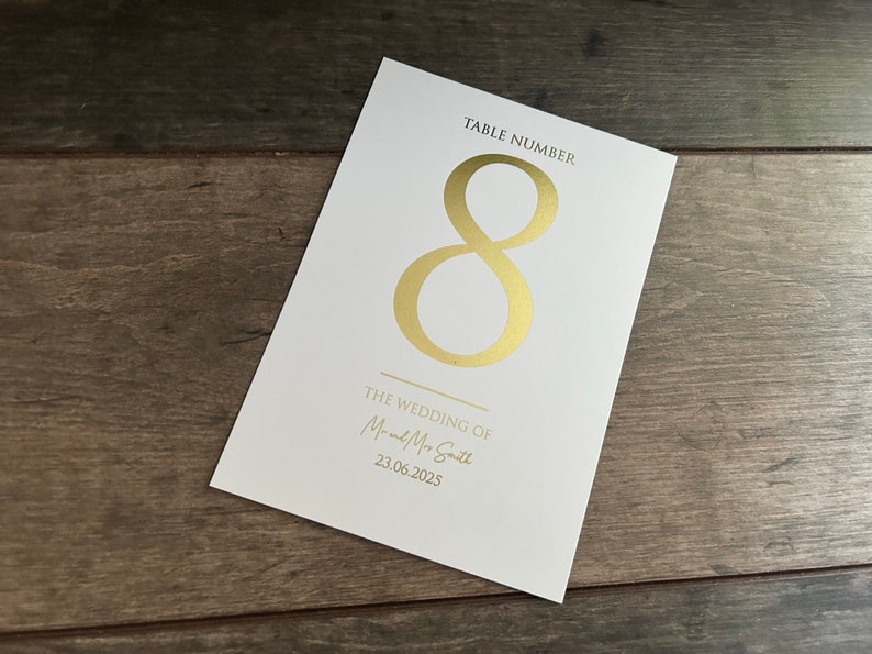 May include: White card with gold foil lettering. The card says "Table Number 8" and "The Wedding of Mr. and Mrs. Smith 23.06.2025".