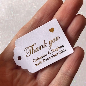 Peut inclure: Etiquette de remerciement blanche avec inscription dorée. L'étiquette dit "Merci" avec un petit cœur doré au-dessus du texte. En dessous du texte, il est écrit "Catherine & Stephen 24 décembre 2021".