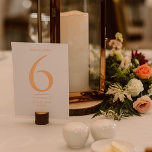 May include: A white card with the text "Table Number 6" in orange and the text "The Wedding of Mr & Mrs Smith" in a smaller font. The card is on a wooden stand and there is a lantern with a candle in the background.