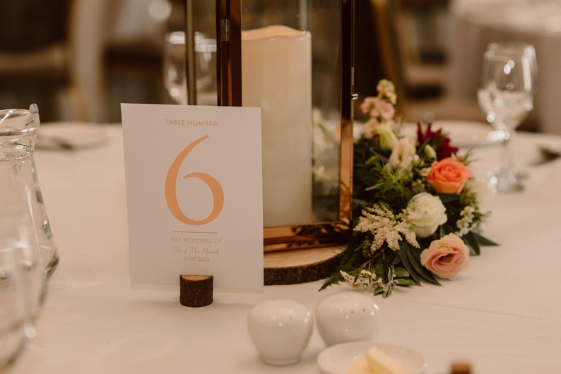 May include: A white card with the text "Table Number 6" in orange and the text "The Wedding of Mr & Mrs Smith" in a smaller font. The card is on a wooden stand and there is a lantern with a candle in the background.