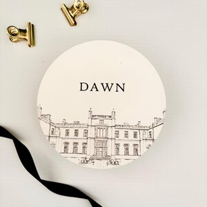 May include: Circular place cards with the word "DAWN" and a detailed architectural illustration. The words "COMING SOON!" are also visible. Two gold binder clips and a black ribbon are also in the image.
