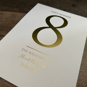 May include: White card with gold foil lettering. The card says "Table Number 8" and "The Wedding of Mr. and Mrs. Smith 23.06.2023".
