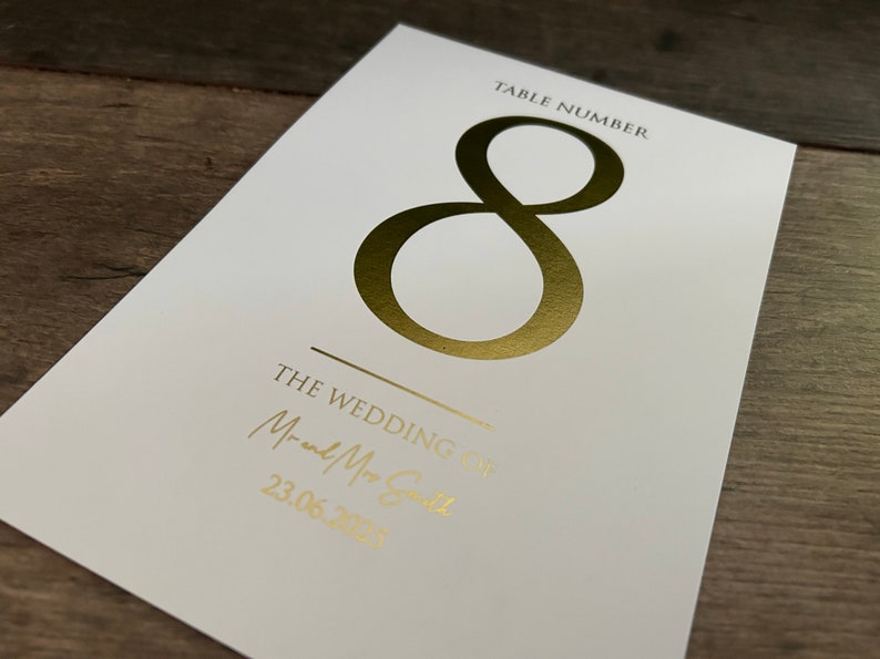 May include: White card with gold foil lettering. The card says "Table Number 8" and "The Wedding of Mr. and Mrs. Smith 23.06.2023".