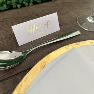May include: White place card with gold lettering that reads "Stephen Smith Groom". The place card is on a wooden table with a gold charger plate and a white napkin.