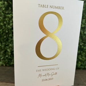 May include: White card with gold foil lettering. The card says "Table Number 8" and "The Wedding of Mr. and Mrs. Smith 23.06.2025".