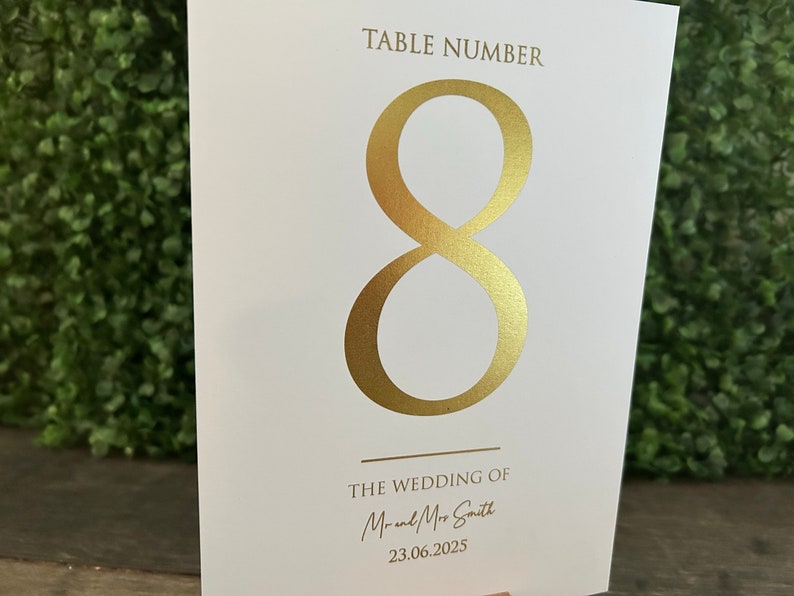 May include: White card with gold foil lettering. The card says "Table Number 8" and "The Wedding of Mr. and Mrs. Smith 23.06.2025".