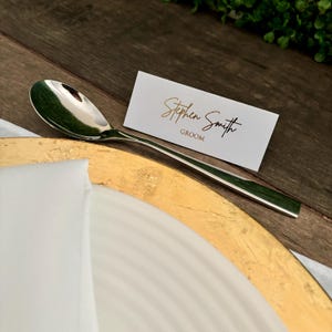 May include: A white place card with gold lettering that reads "Stephen Smith Groom". A silver spoon is placed on a gold charger.