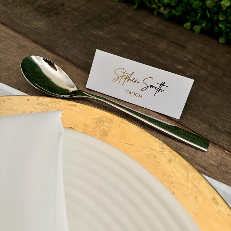 May include: A white place card with gold lettering that reads "Stephen Smith Groom". A silver spoon is placed on a gold charger.