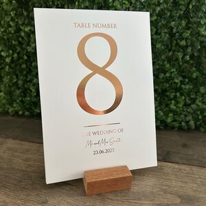May include: White card with a gold foil number 8, the text "Table Number" and "The Wedding of Mr. & Mrs. Smith" and the date "23.06.2023". The card is on a wooden stand.
