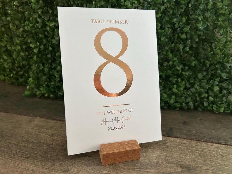 May include: White card with a gold foil number 8, the text "Table Number" and "The Wedding of Mr. & Mrs. Smith" and the date "23.06.2023". The card is on a wooden stand.