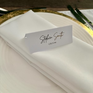 May include: White place card with gold lettering that reads "Stephen Smith Groom".
