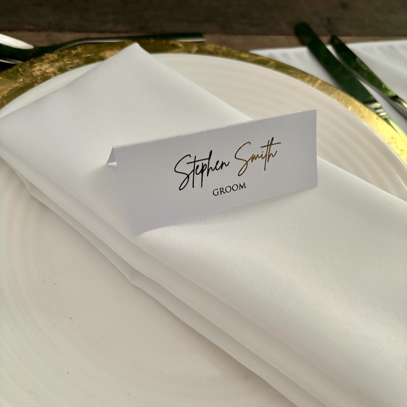May include: White place card with gold lettering that reads "Stephen Smith Groom".