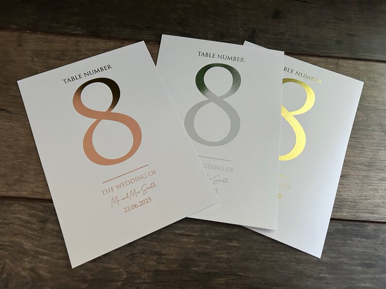 May include: Three table number cards with the number eight printed in gold, silver, and rose gold foil. The cards are white and have the text "Table Number" printed at the top. The rose gold card also has the text "The Wedding of Mr and Mrs Smith 23.06.2025" printed at the bottom.