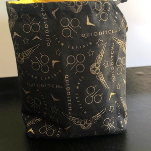 May include: Black drawstring bag with a gold pattern featuring Quidditch-themed graphics, including stars, wings, and goggles. The interior lining is bright yellow. The bag is made of fabric and has a drawstring closure.