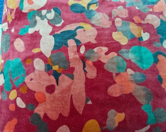 May include: A close-up of a red fabric with an abstract pattern. The pattern features various shapes in teal, orange, yellow, gray, and pink. The fabric appears to be soft and textured, with a slightly wrinkled appearance. The overall design is artistic and colorful.