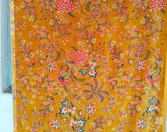 May include: Mustard-coloured fabric with a detailed floral pattern. The design features pink, red, and white flowers with green leaves and stems. The fabric has a soft, velvety texture, and the floral pattern is repeated across the surface.