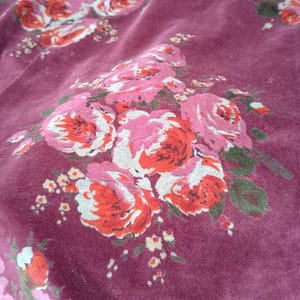 May include: A close-up of a burgundy fabric with a floral pattern of pink and red roses. The fabric has a soft, velvety texture.