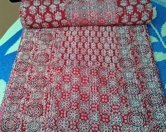 Op de afbeelding: Een opgerolde rode quilt met een herhalend wit en zwart bloemenpatroon. De quilt is gemaakt van een zacht materiaal en wordt waarschijnlijk gebruikt voor beddengoed of decoratie. Het ontwerp is symmetrisch en gedetailleerd.