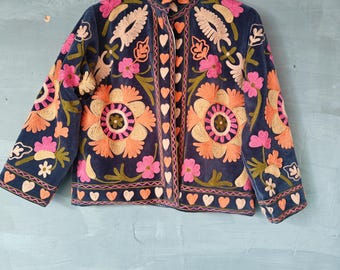Puede incluir: Chaqueta azul marino con intrincados bordados florales en tonos naranja, rosa y beige. La chaqueta presenta un cuello alto y detalles en forma de corazón. Las mangas son de tres cuartos y cuelga de una percha de madera. El estilo es bohemio.