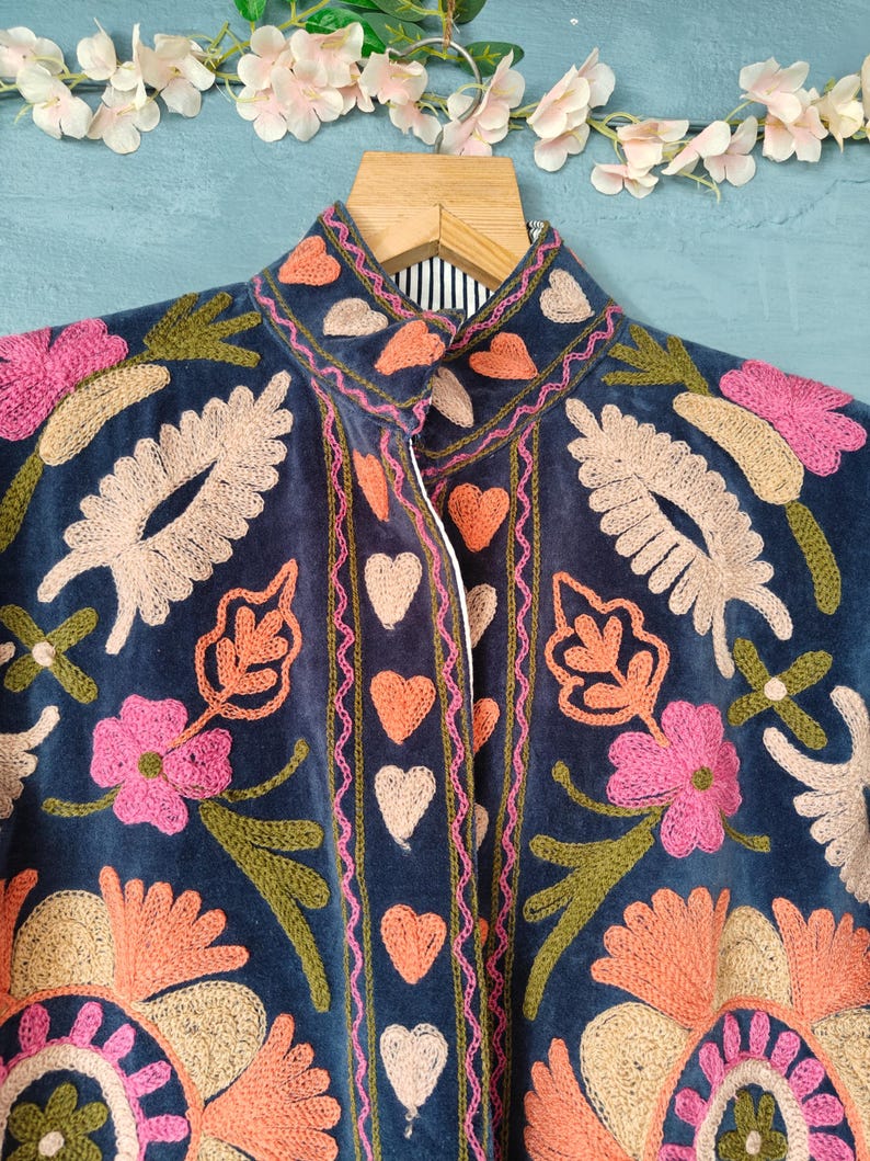 以下が含まれることがあります： ピンク、ピーチ、ベージュの複雑な花の刺繍が施された、ネイビーブルーのベルベットジャケット。 ジャケットはハイカラーで、前面にはハート型の刺繍が施されています。 木製のハンガーにかかっています。