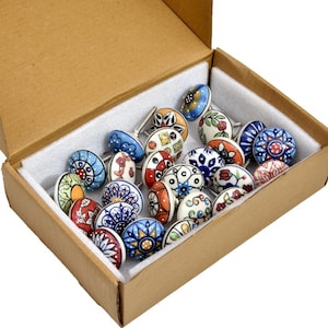 May include: A selection of decorative ceramic cabinet knobs in a cardboard box. The knobs feature hand-painted floral and geometric patterns in a range of colours, including blue, red, and orange. Each knob has a silver metal stem. The box is lined with white foam.