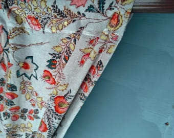 Könnte beinhalten: Cremefarbener Stoff mit floralem Muster in Rot-, Orange-, Gelb- und Grüntönen. Der Stoff ist über einer braunen Stange vor blauem Hintergrund drapiert. Das florale Design zeigt Blätter und Blumen.