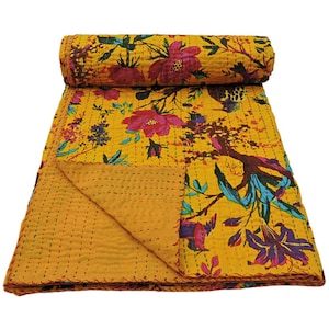 Op de afbeelding: Een gele bloemenquilt met een opgerold gedeelte. De quilt heeft grote rode en roze bloemen, blauwe en paarse accenten en een boomontwerp. De quilt is gemaakt van een zacht materiaal.