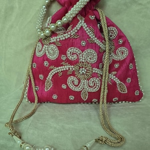 Puede incluir: Un vibrante bolso con cordón rosa con intrincados bordados de perlas y oro. El bolso presenta un asa con cuentas de perlas y borlas decorativas. El diseño incluye patrones florales y de remolino, creando un accesorio elegante y ornamentado.