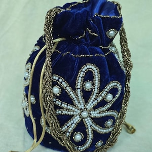 Peut inclure: Un sac en velours bleu marine avec un cordon de serrage et des broderies florales élaborées. Le sac est orné d'un cordon et d'une poignée en perles dorées, et décoré de perles blanches. Le design est symétrique et élégant.