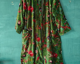 May include: A long, green velvet robe with a floral pattern in red, pink, and purple. The robe has long sleeves and pockets. The interior lining is striped in black and white. The robe is hanging on a wooden hanger.
