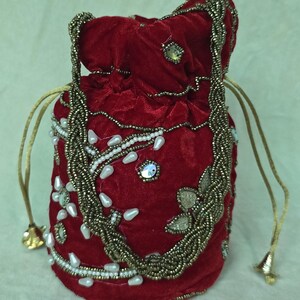 Peut inclure: Un sac rouge en velours avec cordon de serrage, orné de perles et de perles nacrées. Il est doté d'une anse perlée dorée et d'une fermeture à cordon avec des extrémités en forme de cloche dorée. Le motif comprend des perles blanches en forme de goutte et des sequins irisés.