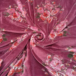May include: Pink velvet fabric with a floral print of pink and red roses. The fabric is folded and draped to show the texture and pattern.