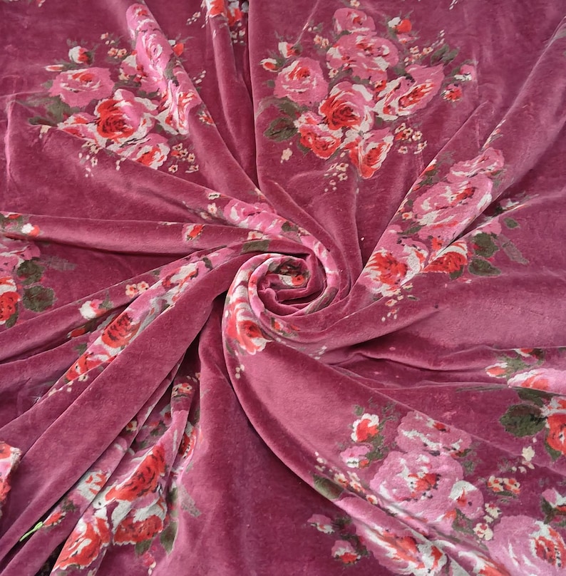 May include: Pink velvet fabric with a floral print of pink and red roses. The fabric is folded and draped to show the texture and pattern.